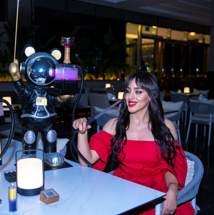 Woman in a red dress enjoying shisha at an outdoor table in a stylish waterfront lounge at night in Business Bay